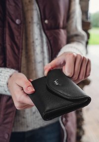 Schwarzes Lederportemonnaie mit abgerundeter Klappe, strukturiertem Oberflächenmaterial und Metalllogo auf der Vorderseite. In den Händen gehalten, mit Fingern, die den Rand greifen.
