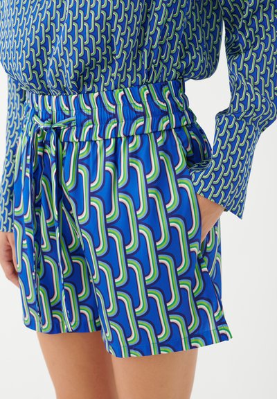 Gros plan d’une personne portant un short bleu à motifs et un haut assorti à manches longues, avec une main dans la poche du short.