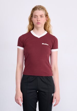 Bordo prilagođena majica s bijelim V-izrezom i akcentima na rukavima, s "Dickies" logotipom u bijeloj boji na prsima. Nosena s crnim hlačama.