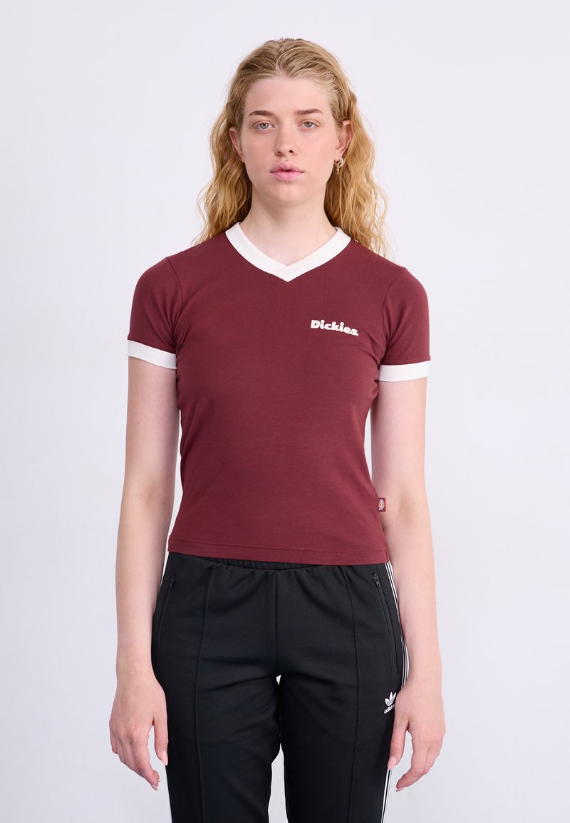 Bordeauxrotes, tailliertes T-Shirt mit weißen V-Ausschnitt und Ärmelausschnitten, mit dem weißen "Dickies"-Logo auf der Brust. Getragen mit schwarzen Hosen.