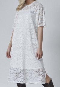 Hvid blondkjole med korte ærmer, blomstermotiv, knælang, med sideslids og broderet logo på brystet. Tekstureret og letvægtsstof.