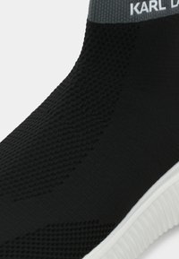 Shoe de malha preta com uma superfície texturada, apresentando um colarinho canelado cinza e o logótipo branco "KARL D." na parte superior. Sola branca.