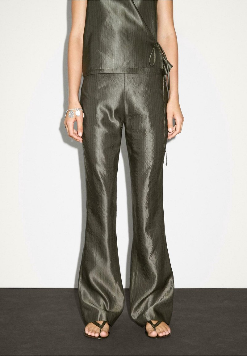 Femme portant un pantalon large brillant vert foncé et un haut portefeuille assorti, debout pieds nus sur un sol sombre contre un mur pâle.