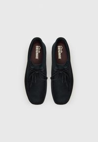 Black suede shoes with a rounded shape, featuring a lace-up design, stitched accents, and a cushioned insoles marked "Clarks Originals."