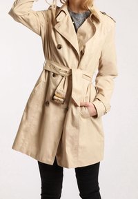 Beige trenchcoat i lättviktsmaterial med dubbelknäppta knappar, bälte i midjan och sidofickor.