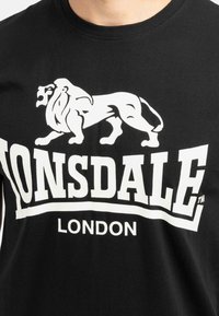 Juodas medvilninis marškinėliai su dideliu balto liūto logotipu ir užrašu "LONSDALE LONDON". Trumpi rankovės ir apvalus iškirpimas.