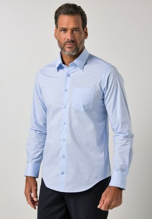 Homme barbu portant une chemise bleu clair à boutons et un pantalon foncé, debout devant un fond gris uni.