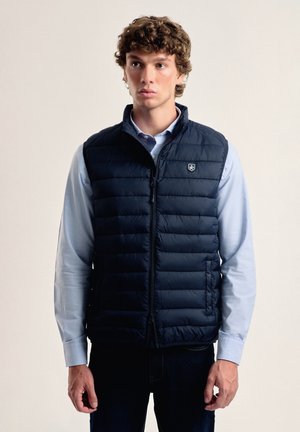 Chaleco acolchado navy con cierre de cremallera, patrón de acolchado horizontal y un logo en el lado izquierdo del pecho. Lleva puesto sobre una camisa de color azul claro.