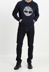 Sudadera con capucha de color azul marino y logo gris, con bolsillo tipo canguro, combinada con jeans oscuros y zapatos negros, vista frontal completa sobre un fondo blanco.