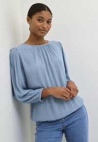 Hellblaues strukturiertes Blusenshirt mit gerafftem Ausschnitt und langen Ärmeln, kombiniert mit enganliegenden Jeans. Schlichte Gestaltung und lässiger Stil.