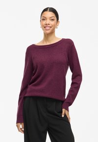 Burgundijas krāsas adīts džemperis ar plašu izgriezumu, garām piedurknēm, ribotām aprocēm un brīvu piegriezumu, savienots ar melniem biksēm.