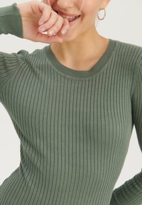 Grüner gerippter Strickpullover mit rundem Ausschnitt und strukturierter Stofftextur. Nahaufnahme der Ärmel und einer Hand des Modells in der Nähe ihres Gesichts.