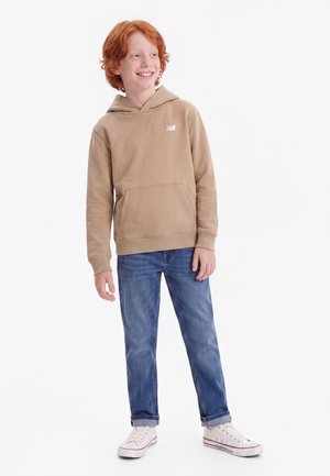 Rødhåret dreng smilende iført en beige hættetrøje, blå cowboybukser med opsmøg og hvide sneakers, stående mod en ensfarvet hvid baggrund.