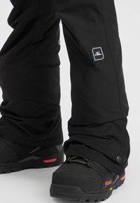 Zwarte sneeuwbroeken met aansluitende manchetten, voorzien van een logo patch en versterkte naden, gecombineerd met zwarte wandelschoenen met rode accenten.