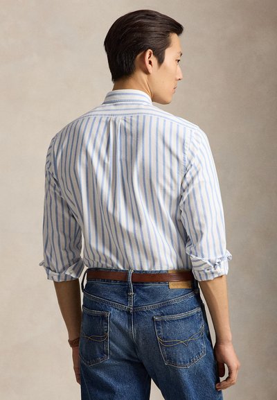 Mann trägt ein blau-weiß gestreiftes Langarmshirt mit hochgekrempelten Ärmeln und blaue Jeans mit braunem Gürtel, von hinten gesehen.