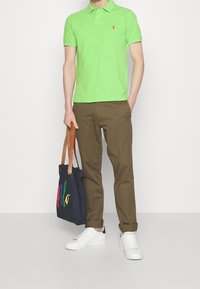 Polo verde con logo rosso, chinos marroni, sneakers bianche e una borsa nera con scritte multicolori. Presentazione di un outfit casual.