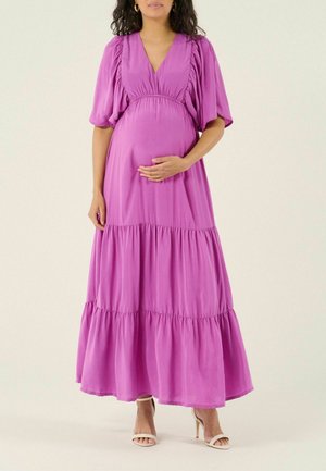 Zwangere vrouw in een vloeiende, gelaagde paarse jurk met pofmouwen en beige sandalen met hoge hakken, die met één hand haar buik vasthoudt.