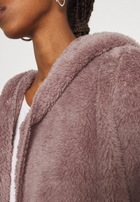 Jas van nepbont in licht mauve met een zachte, gestructureerde afwerking. Voorzien van een capuchon en een open voorkant, gecombineerd met een wit shirt eronder.