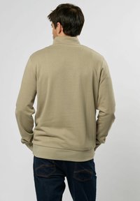 Beige genser med høy krage og ribbede mansjetter, båret over mørke jeans. Materialet ser mykt ut med en jevn tekstur og minimal detaljering.