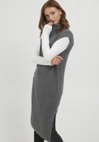 Robe sans manches gris à côtes avec un col haut, comportant une fente sur le côté. Portée par-dessus une chemise blanche à manches longues, assortie à un pantalon noir.