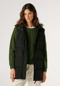 Melnā pūkainā veste ar kapuci un kažokādas apdari, ar rāvējslēdzēju un pogām, valkāta virs zaļa struktūrēta adīta džempera.