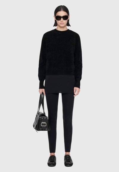Modèle portant des lunettes de soleil noires, un pull noir oversize, un legging et des baskets, tenant un sac à main noir sur un fond gris clair.