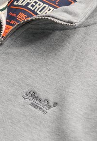 Sweat-shirt à zip gris avec logo "Superdry" brodé et motif orange et bleu partiellement visible en dessous.