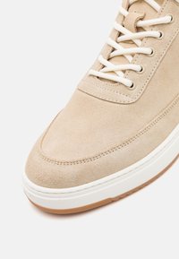 Baskets en daim beige avec des lacets blancs et une semelle blanche épaisse avec une semelle en caoutchouc marron, montrées en gros plan sous un angle.