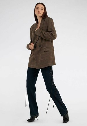 Frau steht vor einfarbigem Hintergrund, trägt ein braunes kariertes, zweireihiges Sakko, schwarze Schnürhose und schwarze Stiefeletten mit Absatz.