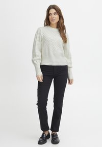 Pull en gris clair texturé avec un motif géométrique, ourlet côtelé, et manches volumineuses associé à un pantalon noir ajusté et des chaussures.