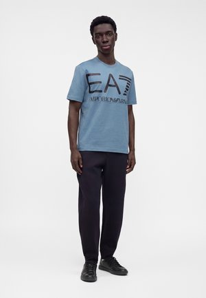 Jeune homme portant un t-shirt bleu clair EA7 Emporio Armani, un pantalon noir et des baskets noires, debout devant un fond blanc uni.