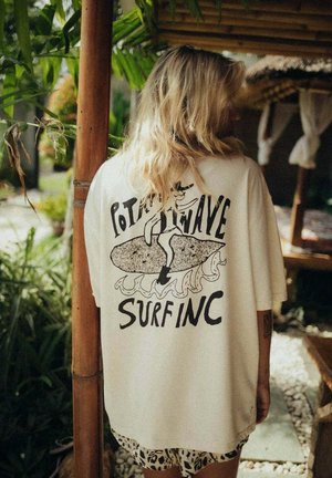 Biała oversize koszulka z nadrukiem grafiki przedstawiającej surfera na fali oraz napisem "POTATO WAVE SURFING" oraz krótkie, wzorzyste spodenki.