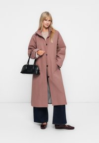 Femme portant un manteau long mauve, un jean large noir, des mocassins bordeaux et portant un sac à main noir sur fond blanc uni.