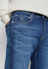 Dunkelblaue Jeans aus Denim mit klassischem Schnitt, versehen mit goldenen Nähten und einem kleinen CK-Logo auf der Vorderasche. Standard-Fünf-Taschen-Design.
