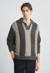 Kötött pulóver galléros nyakkivág�ással, váltakozó sötétzöld és bézs texturált csíkokkal. Két fából készült gomb a nyakkivágás közelében.