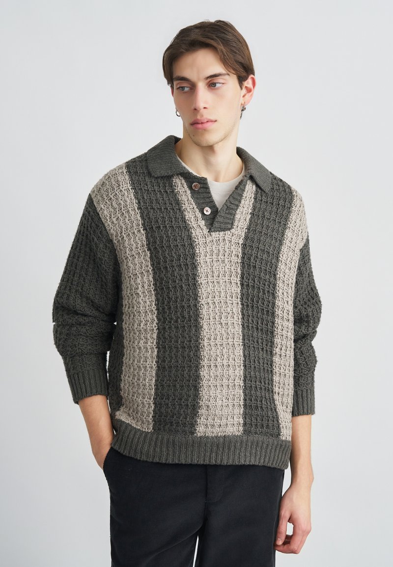 Kötött pulóver galléros nyakkivágással, váltakozó sötétzöld és bézs texturált csíkokkal. Két fából készült gomb a nyakkivágás közelében.