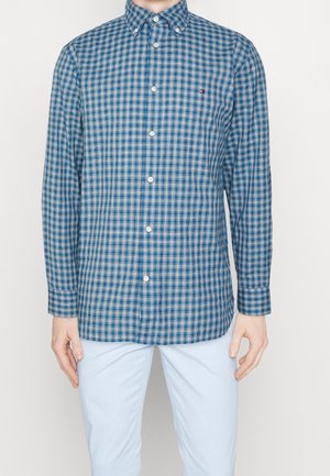 Mężczyzna w niebiesko-białej koszuli w kratę na guziki i jasnoniebieskich spodniach, stojący na tle jednolitego białego tła.