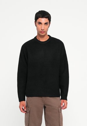 Schwarzer Strickpullover mit rundem Halsausschnitt, lockerem Schnitt und langen Ärmeln. Der Stoff hat eine weiche, strukturierte Optik. Kombiniert mit hellbraunen Cargo-Hosen.