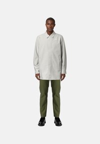 Cappotto grigio chiaro con colletto, dotato di chiusura frontale e tasche laterali, abbinato a pantaloni verde oliva e scarpe nere.
