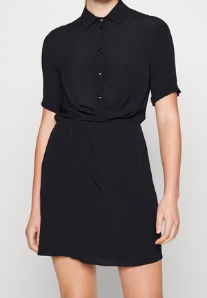 Žena v krátkých rukávech, černých košilových šatech s límečkem, přiléhavým pasem a sukní nad kolena, stojící před jednoduchým pozadím.