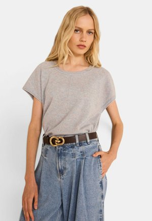 Donna bionda che indossa una maglietta a maniche corte grigio chiaro infilata in jeans blu larghi a vita alta con cintura marrone e fibbia dorata.