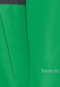 Detailní záběr na zelenou látku s vertikálním švem a okrajem kapsy, s bílým tištěným logem "TEXAPORE" v dolním pravém rohu.