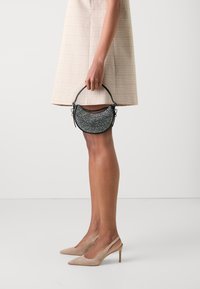 Bolso de mano negro en forma de media luna con embellishments texturizados, pequeña asa y acentos metálicos. Llevado con una falda a cuadros beige y tacones nude.