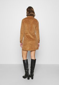 Brun manchesterklänning med långa ärmar, krage och knäppta manschetter. Tillsammans med knähöga svarta stövlar med klack. Slät textur.