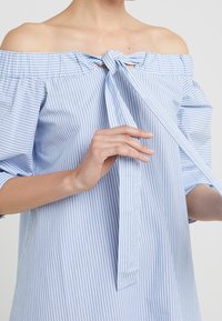 Světle modrá a bílá pruhovaná blůza s odhalenými rameny, krátkými nabíranými rukávy, předním zavazováním mašlí a měkkou, texturovanou tkaninou.