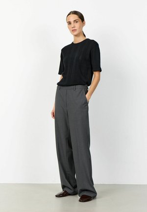 Ženska stoji z rokami v žepih, oblečena v črno pleteno majico s kratkimi rokavi, široke sive hlače in temno rjave čevlje na sivi tlaki.