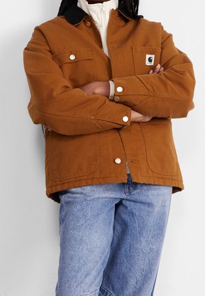 Personne portant une veste marron à boutons avec un col noir et un logo visible, un sweat à capuche blanc zippé en dessous, et un jean bleu clair ample, bras croisés.