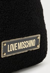 Črn teksturiran material s pravokotno črno in zlato oznako z napisom "LOVE MOSCHINO", pritrjeno z najboljšimi štirimi zlatimi zakovkami.