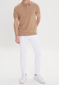 Polo beige avec trois boutons et un col côtelé, associé à un pantalon blanc ajusté et des baskets blanches. Texture de tissu lisse.