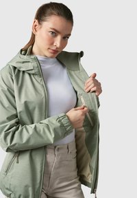 Veste zippée vert clair avec capuche, dotée de poignets élastiques et d'une poche zippée intérieure. Portée par-dessus un haut blanc à la texture lisse.